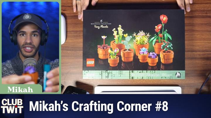 This is a photograph featuring Mikah Sargent holding a Lego "Tiny Plants" set box. The box, with a colorful illustration of potted plants, is placed on a wooden surface. Mikah, wearing a gray fleece and black headphones, is shown from the waist up in a close-up on the left. The background includes a microphone and a blue screen. The image has a watermark reading "CLUB TWIT" and "Mikah's Crafting Corner #8" at the bottom.
